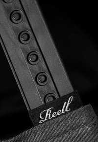 Schwarzer Gurt mit runden Vertiefungen, ausgestattet mit einem Stoffetikett mit weißem Text "Reell" und einem strukturierten grauen Material im Hintergrund.