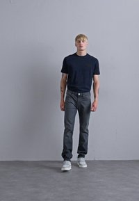 Marinefarbendes T-Shirt mit kurzen Ärmeln, graue Jeans und weiße Sneakers auf einem stehenden männlichen Modell vor einem hellgrauen Hintergrund.