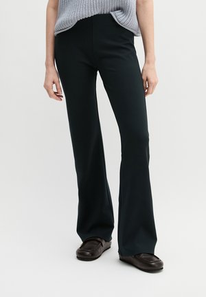 Person trägt schwarze Schlaghosen, schwarze Loafers und ein hellgraues gestricktes ärmelloses Oberteil und steht vor einem weißen Hintergrund.