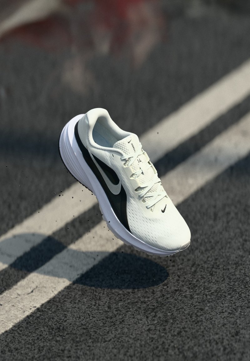 Λευκό αθλητικό παπούτσι Nike με μαύρο λογότυπο swoosh φαίνεται να αιωρείται πάνω από δρόμο με άσφαλτο και λευκές διαγραμμίσεις λωρίδων.
