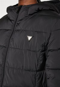 Doudoune noire avec capuche, présentant des sections matelassées, une fermeture éclair à l'avant et un accent de logo triangulaire sur la poitrine. Matériau lisse et durable.