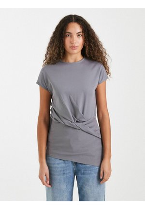 Mujer con el pelo rizado que lleva una camiseta gris de manga corta con detalle retorcido y vaqueros azules, de pie contra un fondo blanco liso.
