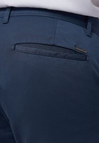 Marineblaue Hose aus glattem Stoff, mit einer horizontalen Gesäßtasche und einem kleinen metallischen Markenakzent am Taschensaum.