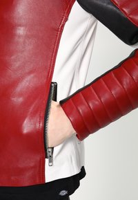Rode en witte leren jas met geborduurde mouwen, zijzak met rits en zwarte accenten. Heeft een aansluitend ontwerp en een soepele textuur.