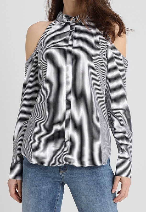 Femme portant une chemise à épaules dénudées à rayures verticales noires et blanches avec des manches longues et un jean bleu, debout devant un fond uni.