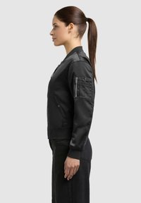 Femme avec queue de cheval brune portant une veste bomber noire et un pantalon noir, debout de profil sur un fond clair uni.