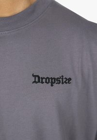 Graues Baumwoll-T-Shirt mit Rundhalsausschnitt und kurzen Ärmeln, versehen mit schwarzem gesticktem Text, der "Dropsize" in einer dekorativen Schriftart zeigt.