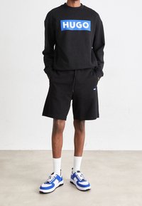 Sweat-shirt noir avec le logo bleu "HUGO", accompagné de shorts noirs et de baskets avec des accentuations bleues. Présente une coupe décontractée et un style athlétique.