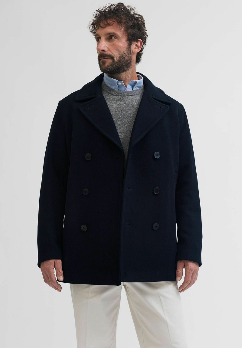 Marineblauwe wollen peacoat met een dubbelzijdig ontwerp, grote zwarte knopen, brede revers en een relaxte pasvorm. Gecombineerd met een grijze gebreide trui en witte broek.