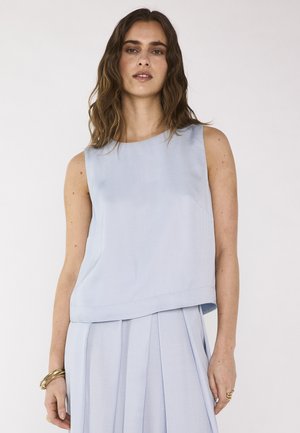 Donna che indossa un top azzurro senza maniche e una gonna plissettata abbinata, con braccialetti d'oro al polso sinistro e anelli sulla mano destra.