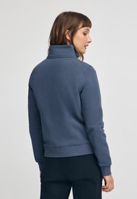 Mörkblå, långärmad sweatshirt med hög krage, ribbade manschetter och figurnära midja. Mjuk textur utan synliga mönster eller detaljer.