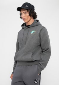 Jovem vestindo um hoodie escuro da Nike Air, calças de moletom cinzas da Nike e um boné preto da Nike, de pé com as mãos nos bolsos.