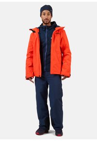 Giacca impermeabile arancione con cappuccio e dettagli neri, indossata sopra una giacca interna isolante blu navy, abbinata a pantaloni blu navy e scarpe nere.
