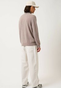 Giovane che indossa un maglione beige, pantaloni bianchi larghi, sneaker bianche e grigie e un cappellino color crema, in piedi di spalle su uno sfondo bianco.