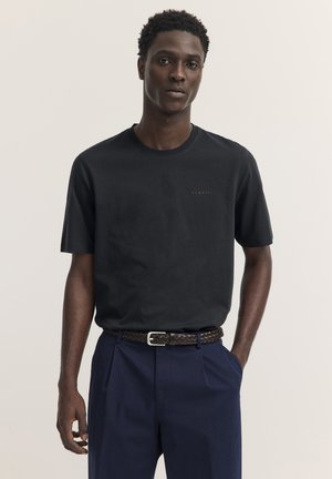 Mann trägt ein schlichtes schwarzes T-Shirt, marineblaue Hose und einen braunen geflochtenen Gürtel, steht mit einer Hand in der Tasche vor einem schlichten Hintergrund.