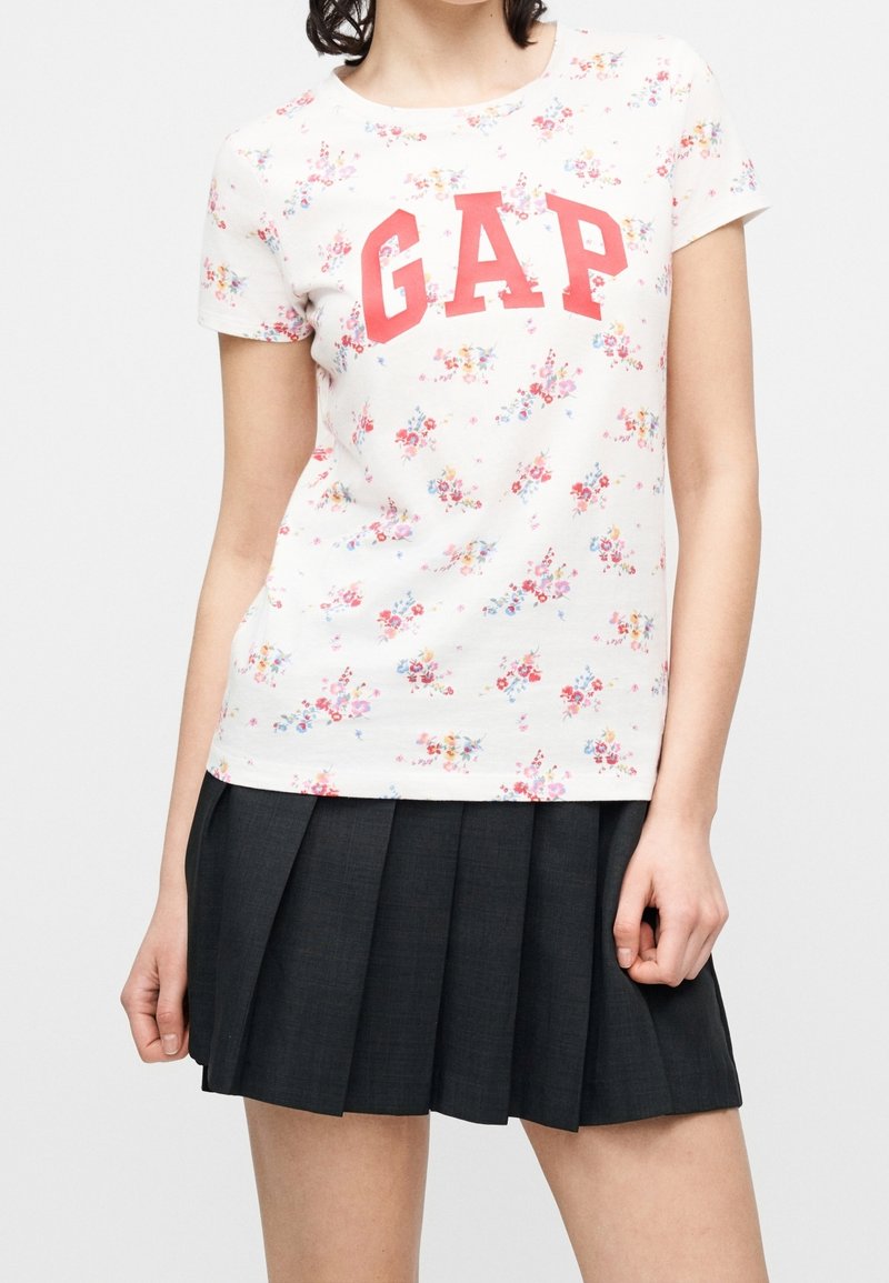 Femme portant un haut blanc fleuri à manches courtes avec des lettres rouges "GAP" et une jupe plissée noire, debout devant un fond uni.