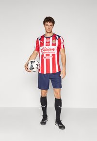 Rød- og hvitstripet fotballskjorte med logo, marineblå shorts, svarte sokker og sko. Holder en svart og hvit mønstrete fotball.