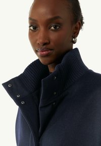 Marineblaues Jacket mit geripptem Stehkragen, Druckknöpfen und glatter Textur. Nahaufnahme, die Material- und Designdetails hervorhebt.