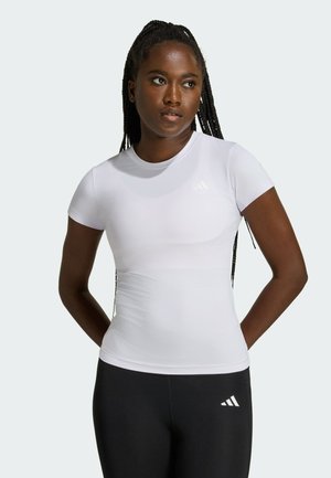 Mujer con el cabello trenzado que lleva una camiseta blanca ajustada de manga corta y leggings negros con un logo blanco, de pie frente a un fondo sencillo.