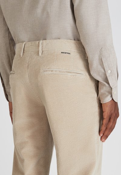 Beige corduroy broek met prominente verticale ribbels, voorzien van een achterzak en een logo detail. Een gladde, gestructureerde afwerking is zichtbaar.