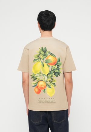 Mann trägt ein beiges T-Shirt mit großem buntem Aufdruck von Zitronen, Orangen und grünen Blättern auf dem Rücken, kombiniert mit dunklen Jeans.