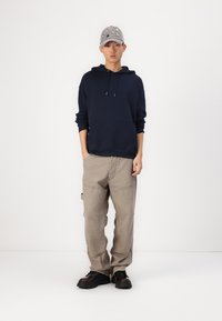 Marine hettegenser, beige bukser, grå caps med broderte detaljer. Svarte sko med teksturerte såler. Ensfarget bakgrunn.