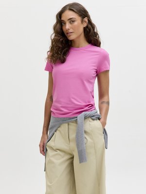 Vrouw met lang krullend haar, gekleed in een roze T-shirt, beige wijde broek en een grijze trui om haar middel geknoopt.