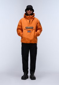 Orange waterproof jacket with a front pocket, logo patch, and Norwegian flag. Paired with black cargo pants and black footwear, worn with sunglasses.