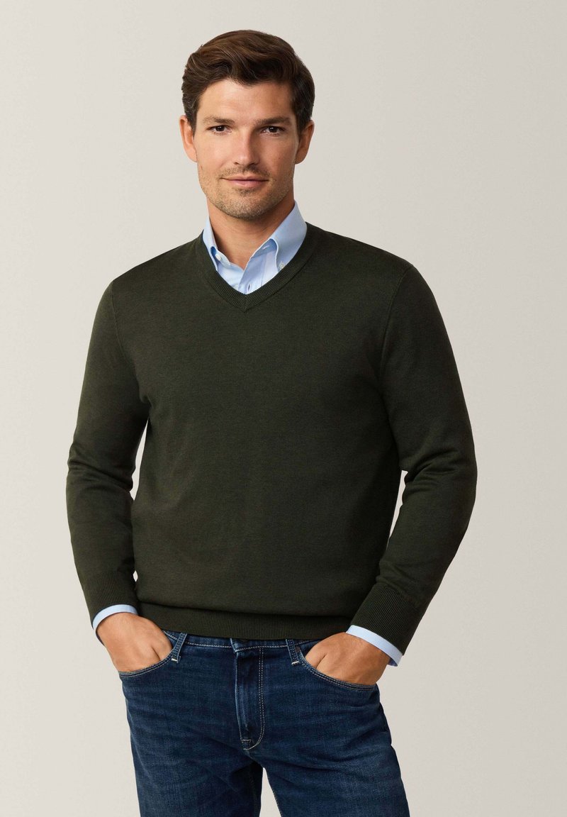Suéter verde oscuro de cuello en V en un material de punto suave, llevado sobre una camisa de collar azul claro, combinado con jeans oscuros. Diseño simple y ajustado.