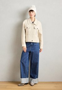 Beige lühike jakk kahe esipocketiga, koos laiadest jänesepüksidest, millel on purustatud servad, ja valge kudumise beanie'iga.