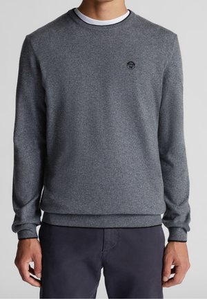 Ein Mann trägt einen grauen Langarmpullover mit rundem Ausschnitt und kleinem schwarzem Logo auf der linken Brust sowie eine dunkle Hose und steht vor einem schlichten Hintergrund.