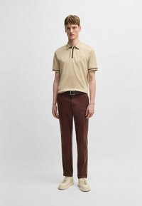 Beige polo shirt with brown and cream accent stripes, paired with brown trousers and cream sneakers. Model stands in front of a plain background.
