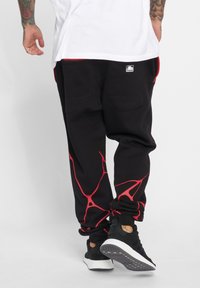 Schwarze Sweatpants mit roten abstrakten Mustern an den Seiten, ausgestattet mit zwei Gesäßtaschen und einem lockeren Schnitt. Kombiniert mit schwarzen Sportschuhen.