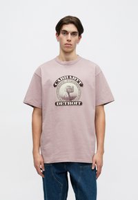 Svetlo roza bombažna majica s krožno grafiko, ki vsebuje besedila "Carhartt WIP" in "Detroit" v temno rjavi barvi. Okrogla izreza, kratki rokavi.