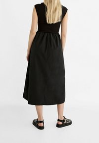 Vestito maxi nero con corpetto aderente e gonna ampia e arricciata. Maniche corte e texture liscia e opaca. Indossato con sandali neri flat.