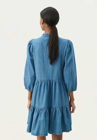 Denim suknelė su susirinkusiu dizainu, pūstomis rankovėmis ir sluoksniuota sijoną. Audinys yra vidutinio mėlynumo, sklandžios tekstūros ir elastingais rankogaliais.