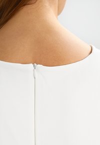 Weißes Kleidungsstück mit glattem, strukturiertem Stoff und einem Reißverschluss am Nacken. Einfaches, sauberes Design ohne Muster.