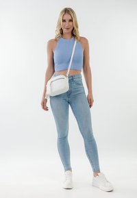 Lyshåret kvinde iført blå ærmeløs cropped top, lyseblå skinny jeans, hvide sneakers og en hvid skuldertaske, stående på hvid baggrund.