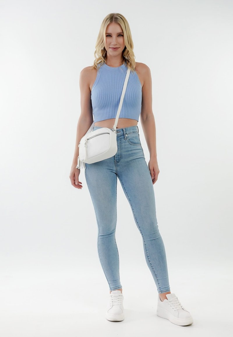 Lyshåret kvinde iført blå ærmeløs cropped top, lyseblå skinny jeans, hvide sneakers og en hvid skuldertaske, stående på hvid baggrund.