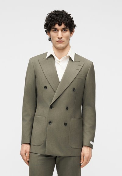 Άνδρας με σγουρά μαλλιά, ντυμένος με διπλό σακάκι ελιάς και λευκό πουκάμισο με γιακά, στέκεται μπροστά σε απλό φωτεινό φόντο.