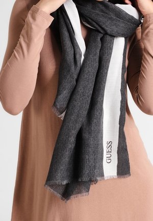 Personne portant un haut à manches longues marron avec une écharpe gris foncé et blanche autour du cou, comportant le nom de la marque "GUESS".