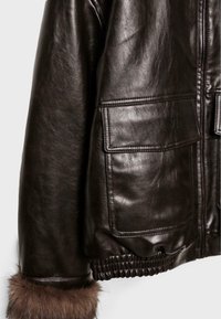 Giacca in pelle marrone con finitura lucida, dotata di due tasche frontali, polsini foderati con dettagli in pelliccia e orlo raccolto in vita.