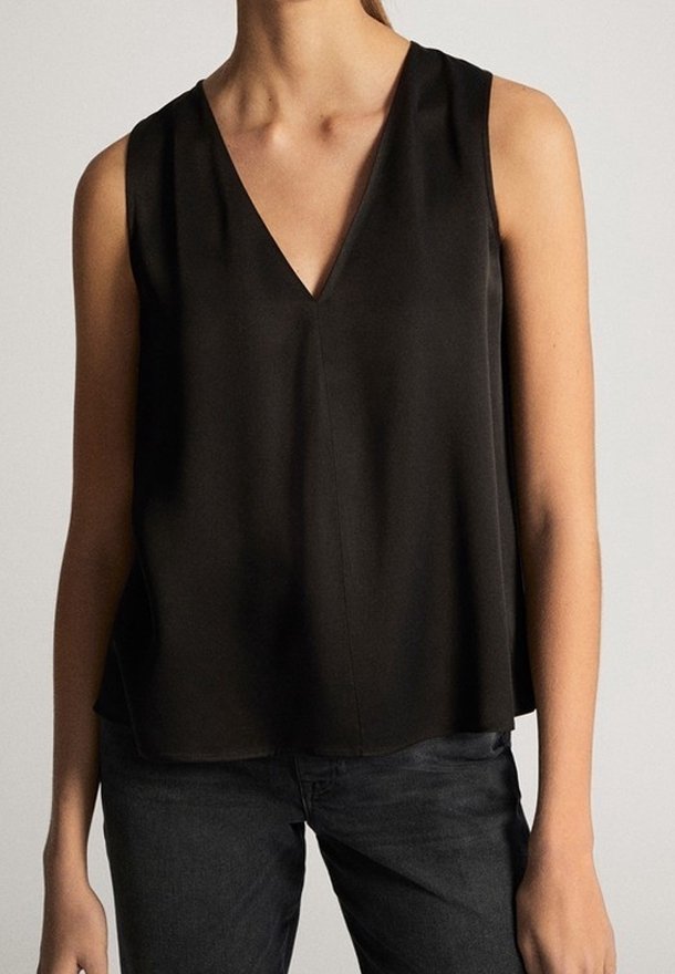 Femme portant un haut noir sans manches en col V et un jean foncé, debout devant un fond neutre.