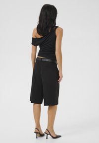 Black sleeveless top with draped fabric and high waist, paired with black knee-length tailored shorts and black heeled shoes.