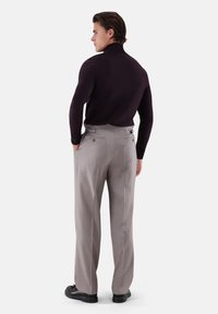 Mann trägt dunklen Rollkragenpullover und hellgraue, hoch taillierte Hose mit Gesäßtaschen, steht mit teilweise in die Taschen gesteckten Händen, Rücken zur Kamera gewandt.