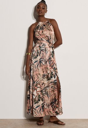 Femme portant une robe maxi sans manches à col halter avec un imprimé feuilles tropicales rose, vert et beige, debout contre un mur uni.