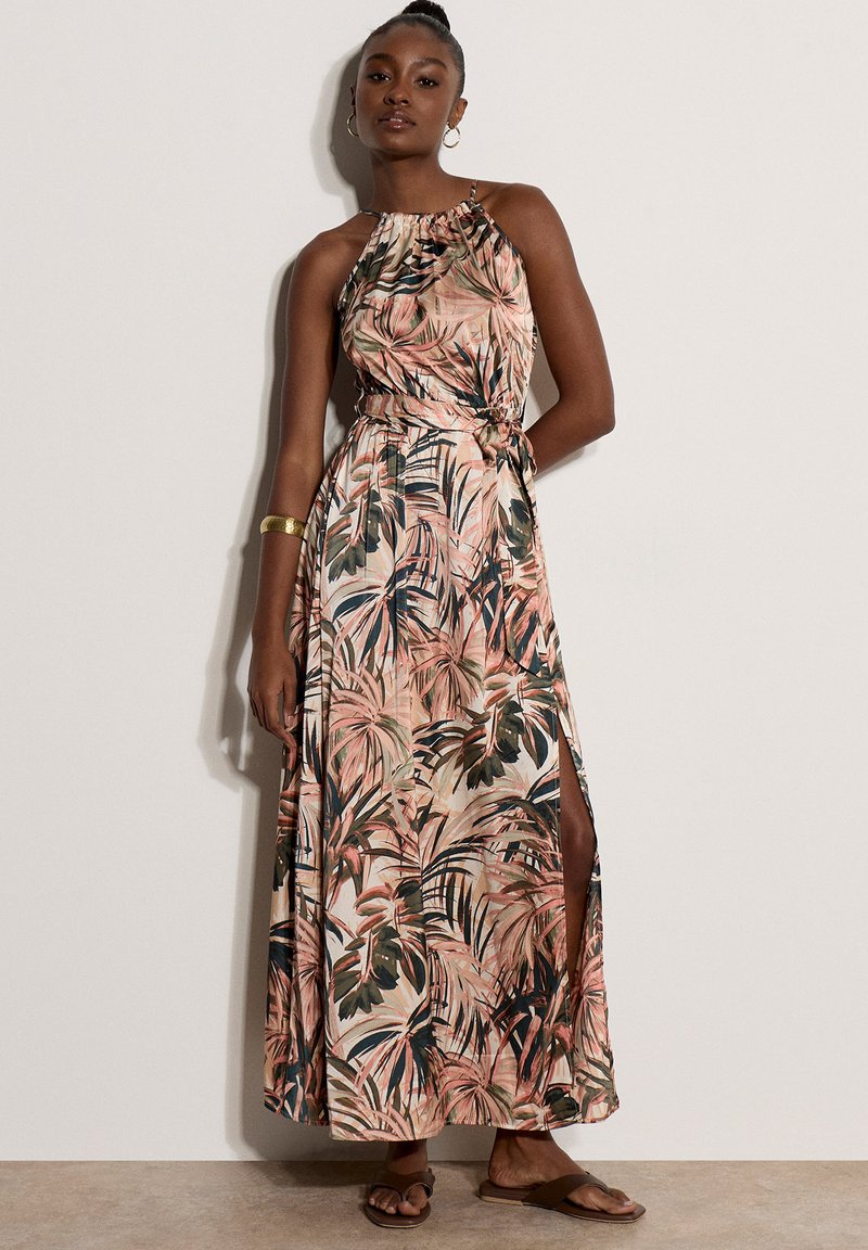 Femme portant une robe maxi sans manches à col halter avec un imprimé feuilles tropicales rose, vert et beige, debout contre un mur uni.