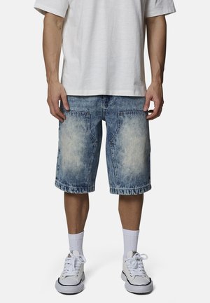 Homme portant un short en jean bleu délavé, des chaussettes blanches montantes, des baskets blanches et un t-shirt blanc uni, debout devant un fond blanc.