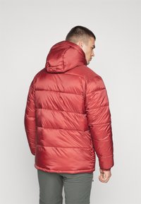 Jachetă puf red cu model matlasat orizontal, design cu glugă și un finisaj lucios, asociată cu pantaloni verzi. Fără detalii vizibile de hardwear.