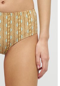 Κοντινό πλάνο στο ισχίο και το χέρι μιας γυναίκας που φοράει μαγιό με ψηλή μέση, σε χρώματα μαυρισμένου και μπλε γεωμετρικά ηλιακά μοτίβα.